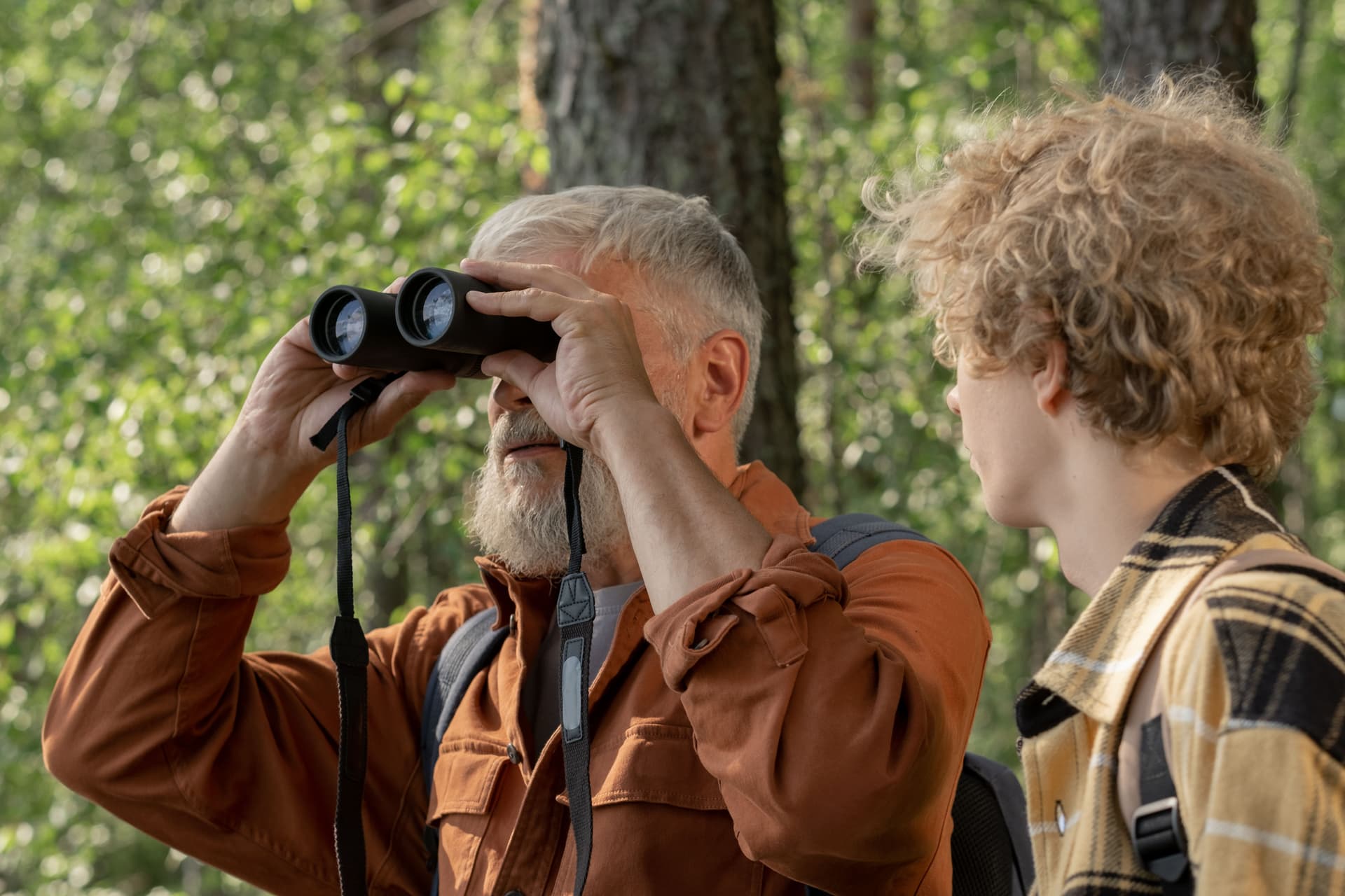 Ein älterer Erwachsener blickt neben einem Kind im Wald durch ein Fernglas; gemeinsame Naturbeobachtung, Ort unbestätigt.
