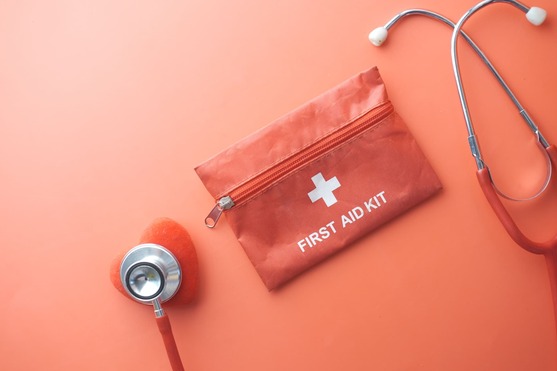 Eine geschlossene rote Erste-Hilfe-Tasche mit weißem Kreuz neben einem Stethoskop auf rosa Untergrund; illustratives Gesundheitsplanungsfoto.