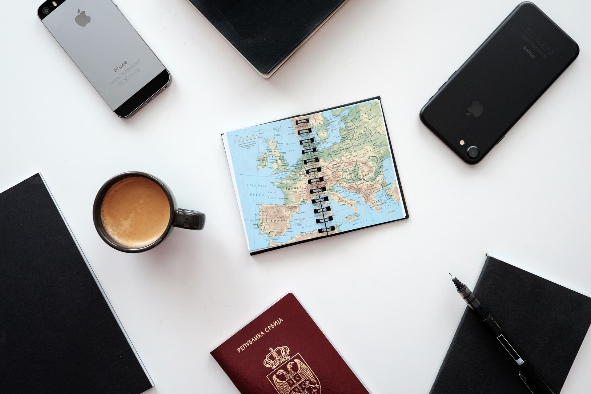 Zwei Smartphones, ein serbischer Reisepass, ein Kartennotizbuch und Kaffee auf einem weißen Tisch; illustratives Reisevorbereitungsfoto.
